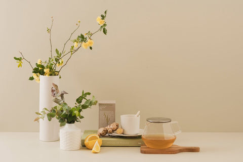 A cup of tea sat on top of a book, with ginger and lemon beside it and a Kinto Carat teapot in front