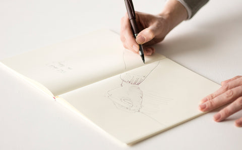 A Midori notebook lays flat on a white surface, with a hand holding a pen hovering over the page and a loose sketch of a person in progress