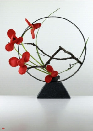 'Tsuboniwa', an ikebana arrangement made with tulip, cordyline leaf and cast iron pot. A slim black ring stands upright on top of the trapezoidal cast iron pot, with a small branch reaching from right to left. Four flowers mark the lower left edge of the circle, stark red against the black metal