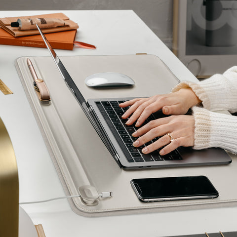An Orbitkey Desk Mat (a rectangular stone-coloured mat laid on a desk) with a laptop, phone, and mouse resting on it as a pair of hands type at the laptop keyboard