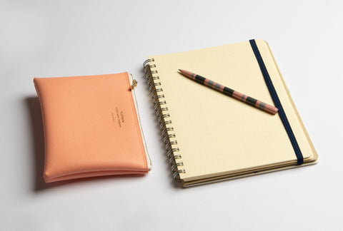 A Delfonics notebook lays open on a grey surface, a pen laid across its exposed Grid page. Next to it, a peach-coloured wallet.