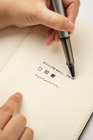 A hand holding a LAMY AL-star rollerball pen and writing "rollerball pen" on a notebook page. Below it are three examples of how a rollerball pen writes.