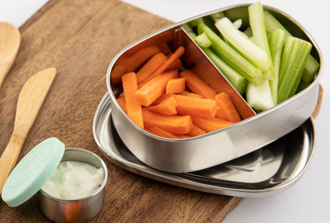 An Ever Eco Stainless Steel Bento Box open, filled with carrot and celery, and laid on top of a chopping board with a small container and bamboo cutlery