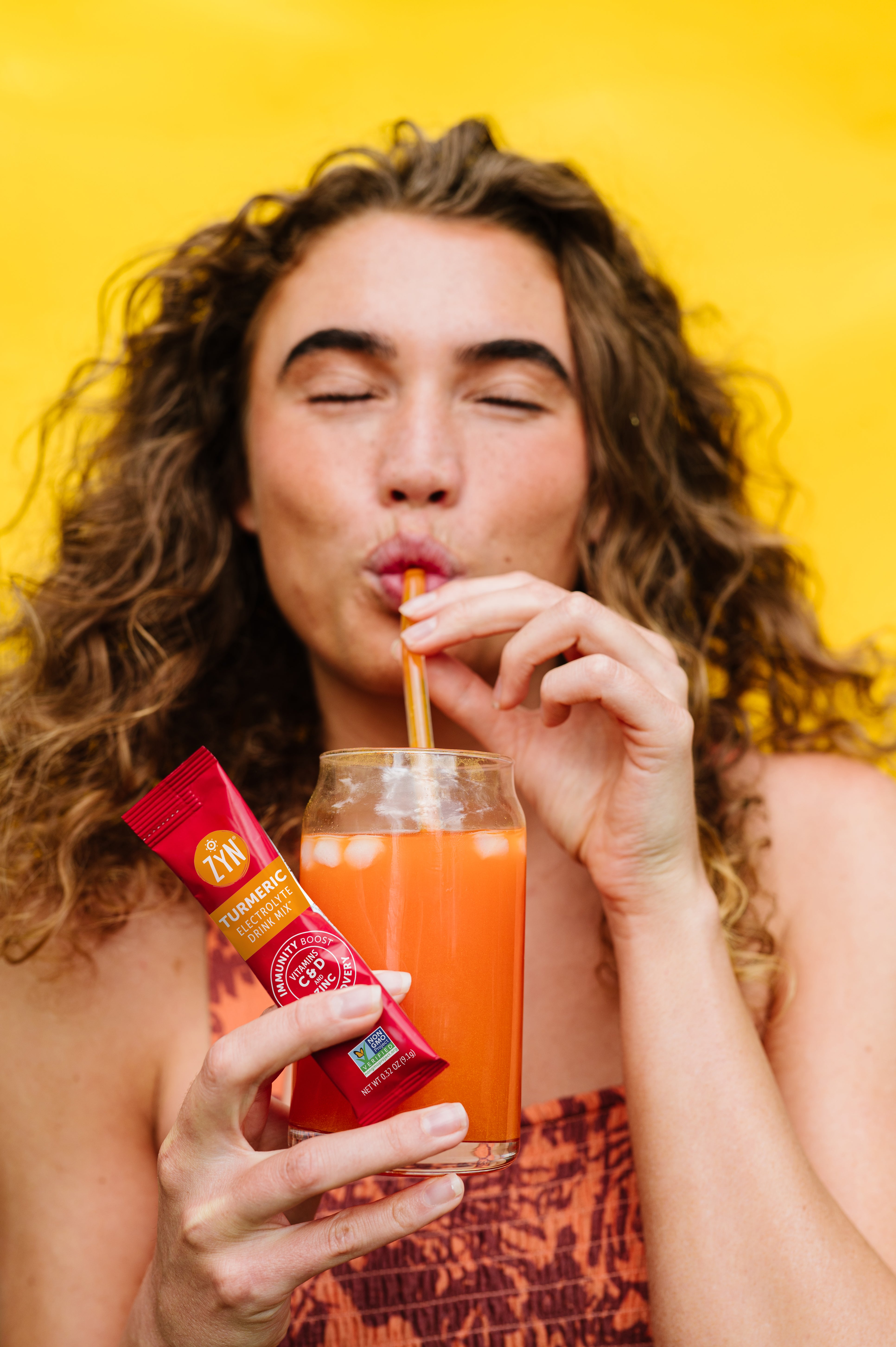 Person sipping orange drink, holding turmeric mix packet against yellow background.