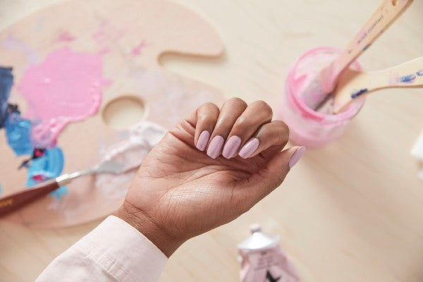 Hand in a loose fist showing baby pink polish over a paint pallet and brush