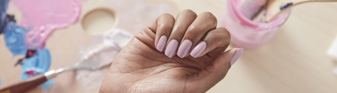 A hand with a loose fist shows mauve nails with paint brushes in the background