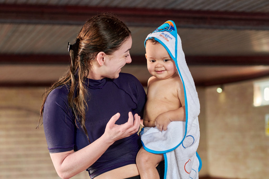 Water Babies teacher holding a little one at poolside. The little one is wearing a hooded baby towel.