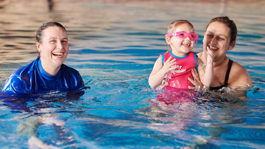 Water Babies teacher smiling, side by side with a carer and their toddler, who is wearing a toddlers wetsuit and pink goggles.