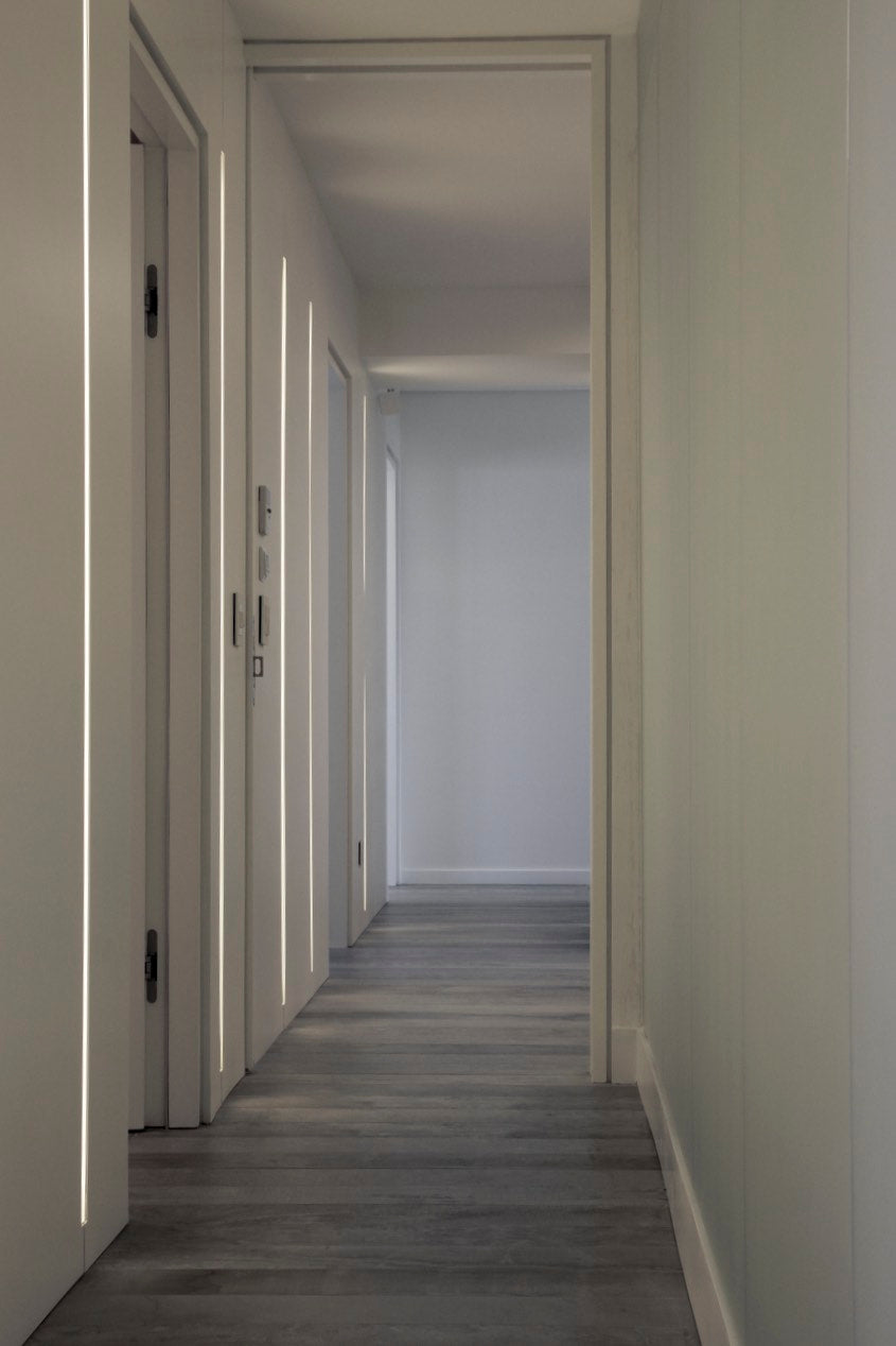 Corridor Design, a minimalist interior design, grey and white, with a hidden wall recessed warm light fixture to contrast smoothly with the light grey wood tile floor.