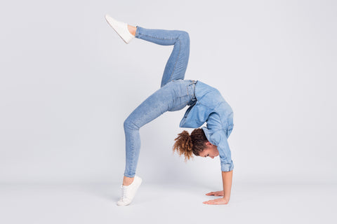 Woman doing a backwards bridge movement with one leg in the air, wearing all denim in front of a grey background