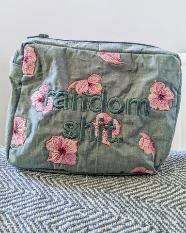 A zipped pouch made with a sage green chambray with large embroidered flowers in pink and the words 'Random Shit' embroidered on it.