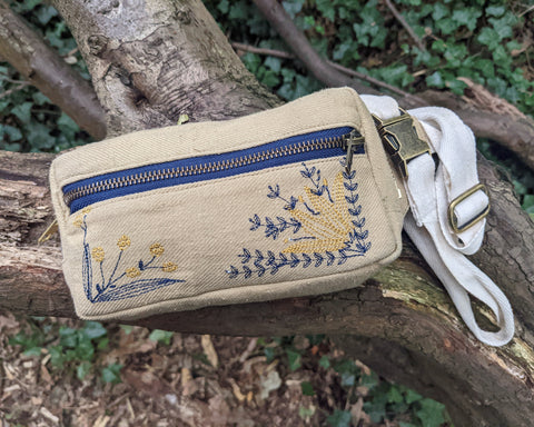 A bum bag in a buff coloured canvas with dark blue zip and blue and yellow embroidered flowers, all photographed in a woodland setting.