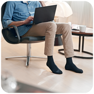 Man sitting in an armchair with a laptop, wearing Sigvaris Essential Cotton compression socks in Black