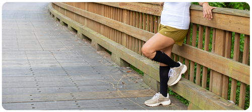 Woman in athletic clothing resting against a railing, wearing Sigvaris Athletic Recovery compression socks in Black