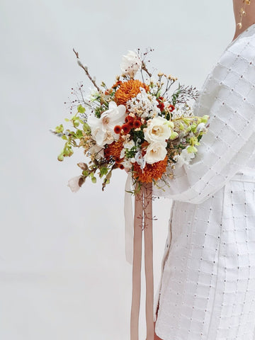 Vrouw met witte jurk houdt modern bruidsboeket vast met roestkleurige en witte tinten en oranje chrysanten, helleborus en phlox.