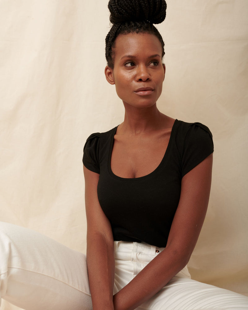 Side profile of black supima cotton tulip sleeve scoopneck tee on model in white jeans sitting on floor and looking to side