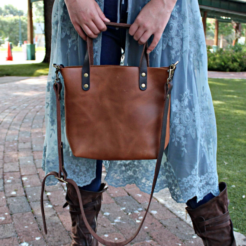 A model on a brick sidewalk holding her full-grain crossbody tote bag by Kerry Noël. In fall fashion, the bag matches her riding boots and cute blue cardigan perfectly.