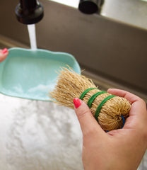 Woman with pink nail polish using an escobeta de raiz Mexican dish scrubber at the sink with water running