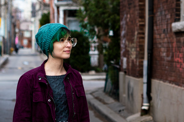 A non-binary white person with teal hair and glasses is standing in a Toronto alley, wearing a purple denim jacket, a grey sweater, and a teal hat.
