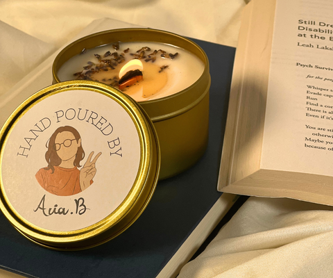 image of Goodness Gift tin candle lit on book on top on blanket. Lid of candle is propped up by candle, with label of associate holding peace sign.