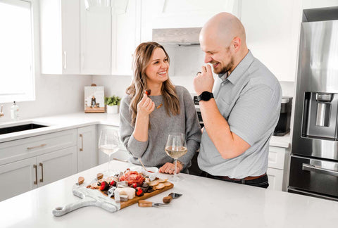 Husband and wife sharing a Charcuterie board of meat and cheese in a white kitchen on a resin accented cheese board