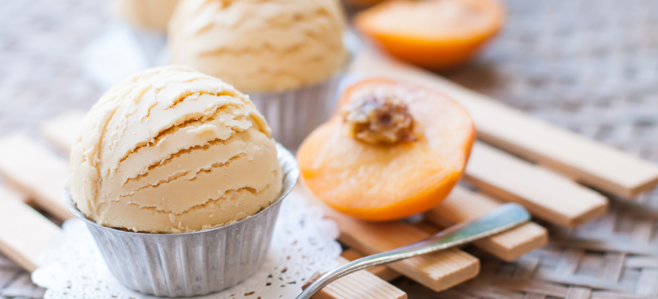 donvier peach ice cream receipe. peach ice cream scooped out into metal baking cups with sliced peach lay on the side on a wooden slat placemat.