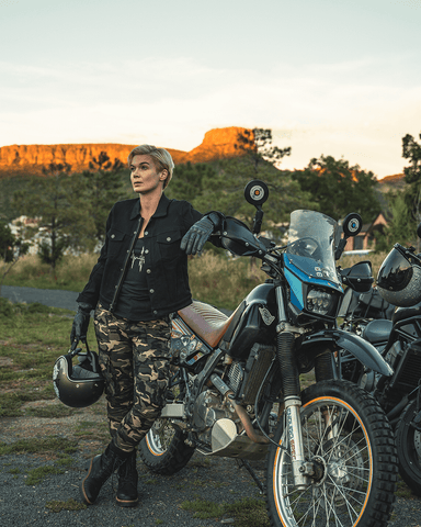 An image of a blonde woman with short hair looking in the distance while leaning on her motorcycle and wearing the Lara Cargo motorcycle pants (women's)