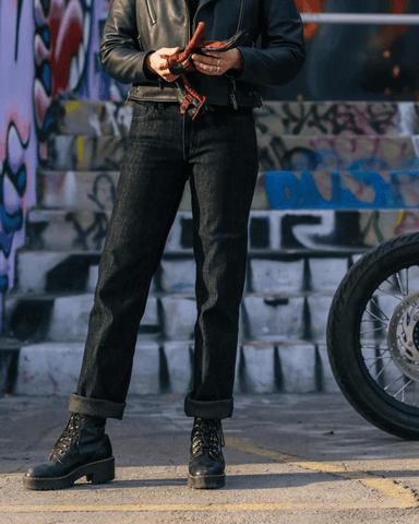 An image of a woman standing in front of a mural wall wearing the Backroads motorcycle jeans (women's)