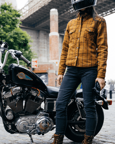 An image of a woman standing in front of her motorcycle in front of the Brooklyn Bridge wearing the Cordura motorcycle jeans for women