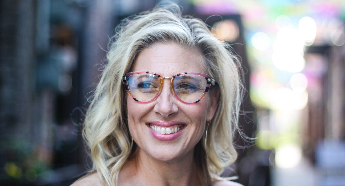 Alt text: headshot of Stormy Hinkle adult woman smiling with stylish pink and black cateye glasses and curled blonde hair