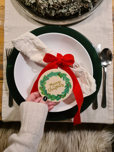 gestalteter Untersetzer mit grünem Kranz und goldener Schrift "Merry Christmas" dekoriert auf einem Teller angelehnt an eine Leinenserviette, mit roter Schleife gebunden