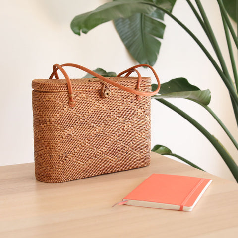 Bali rattan tote bag by Ganapati Crafts Co. is put on a wooden desk with a pink gold notebook next to it