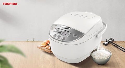 A close-up of a Toshiba rice cooker with 3D IH heating technology, featuring a digital control panel with various cooking options. The rice cooker is white with a silver panel and handle. To the right, there is a small gray bowl filled with steaming white rice, along with a pair of chopsticks resting on a wooden table. The accompanying text mentions '3D IH heating with full heating for great tasting rice,' describing the efficient heat conduction and cooking capabilities of the appliance.