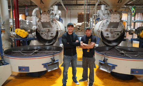 Martin and his colleague Alfredo stand in front of giant coffee roaster machines