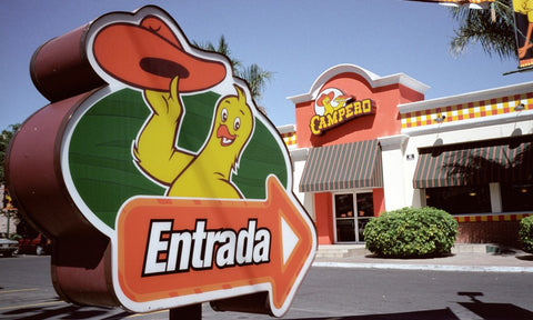 Outside view of a Pollo Campero restaurant, with the welcome sign in the foreground, which features a yellow chicken in a sombrero