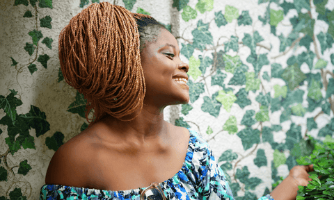An afro-latina woman smiles as she stands against a wall with ivy-covered wallpaper