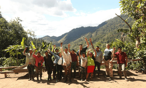 A group of 13 coffee farmers wave in front of a backdrop of verdant hills