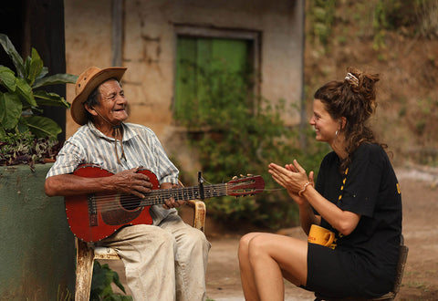 An elderly Latino man sits playing guitar outside his house while a young girl claps along