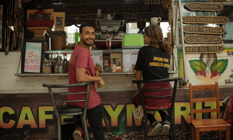 Two people, wearing 'Hecho con Orgullo Latino' shirts sit at a coffee cart in Central America