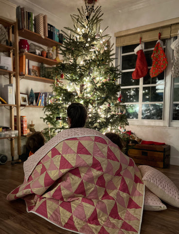family gathered around christmas tree draped in a pink and gold quilt