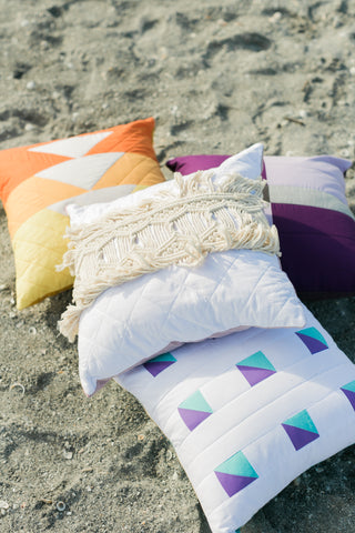 quilted pillow with macrame laying in pile with other quilted pillows on beach