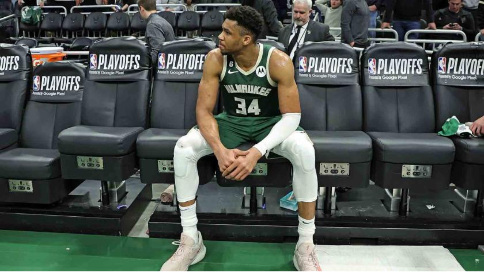 Giannis Antetokounmpo sits in the centre of a row of leather seats with the NBA logo and the word playoffs written on the back. Giannis is wearing his green game jersey and shorts looking off to left.