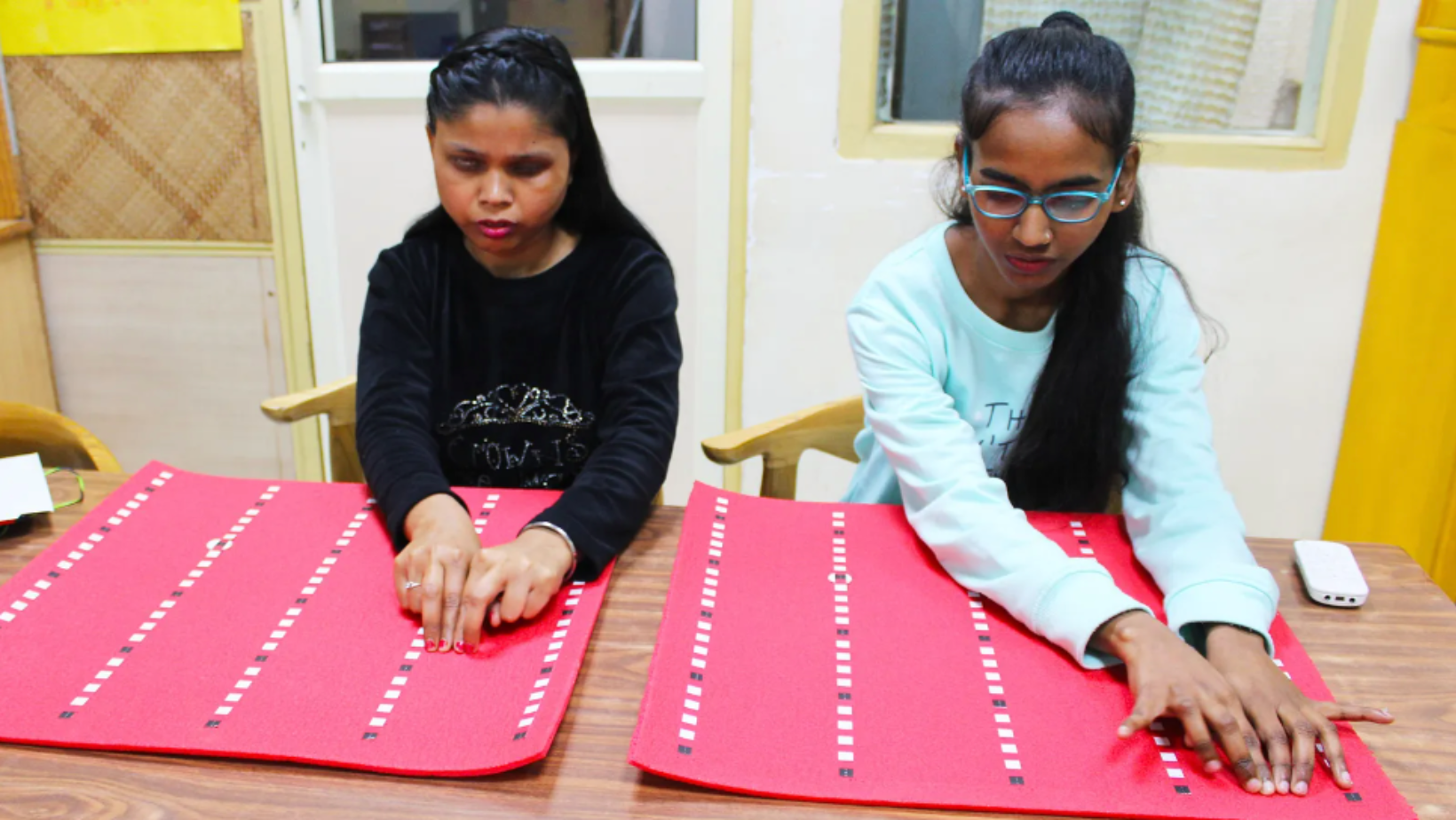 Two Indian women are dressed casually and seated at a table, reading braille on specially designed flexible sheets.