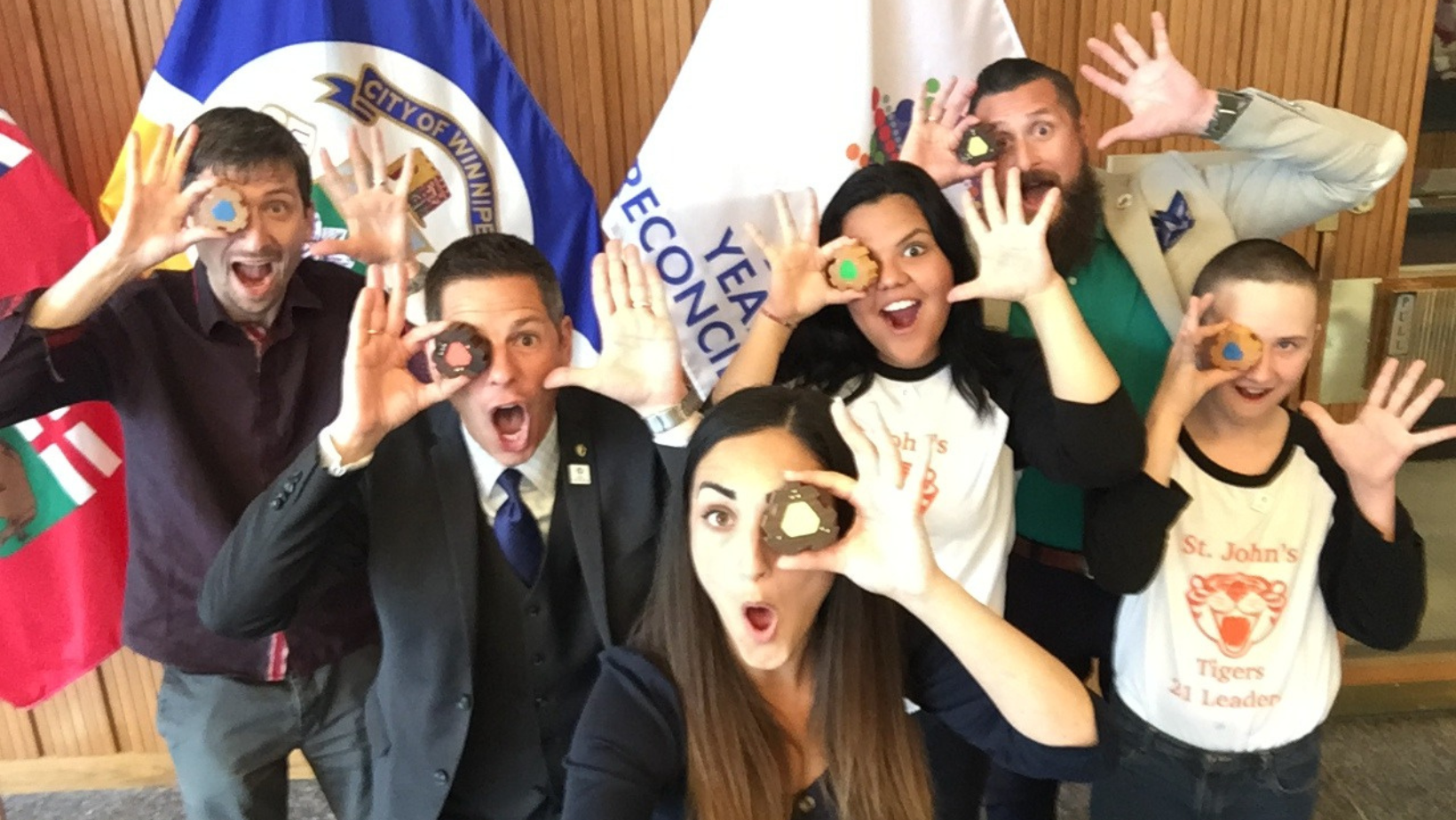 6 people stand in front of a series of flags inside Winnipeg City Hall. The people are standing in sort of a pyramid formation, with Twenty One Toys Founder Ilana Ben-Ari in the centre. On the left side of the picture are Twenty One Toys team member Ryan Burwell and Mayor Brian Bowman. On the right side of the picture is Vice Principal Cree Crowchild and 2 St Johns’ High School students wearing matching black and white shirts.  Each person is holding up a piece of the empathy toy to cover one eye, holding their other hand with the finger stretched out next to the opposite  side of their face, and making silly faces.