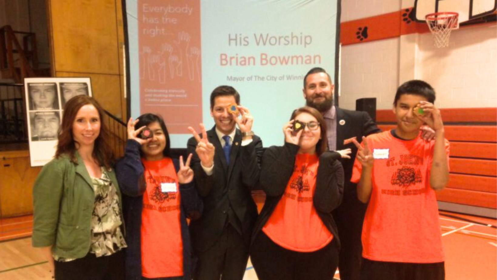 6 people stand in a school gym in front of a projector screen that shows the message His Worship Brian Bowman. The people standing in front are guidance counsellor Robin Wilson, Mayor Brian Bowman, Vice Principal Cree Crowchild, and 3 St Johns’ High School students wearing matching orange t-shirts. The students and the mayor are each holding up a piece of the empathy toy to cover one eye, making a peace sign with their other hand, and smiling.