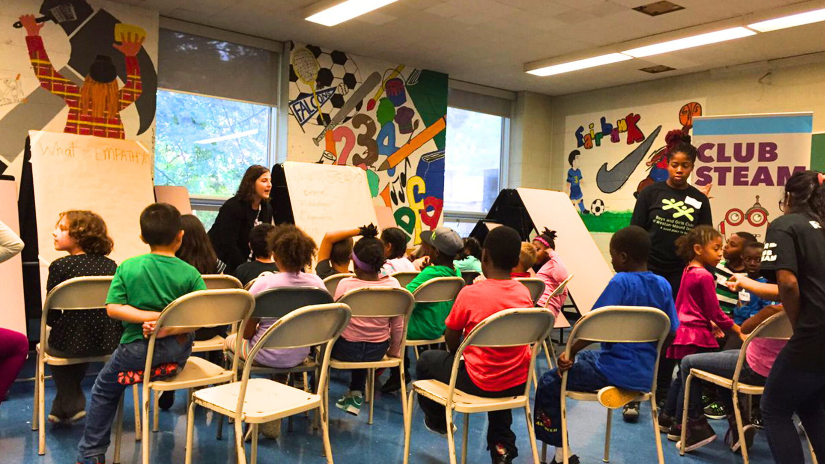 A group of children sits on chairs facing away from the camera. They face couple of flip charts, one of which says What is Empathy at the top. A woman stands next to the flip chart facing the children.