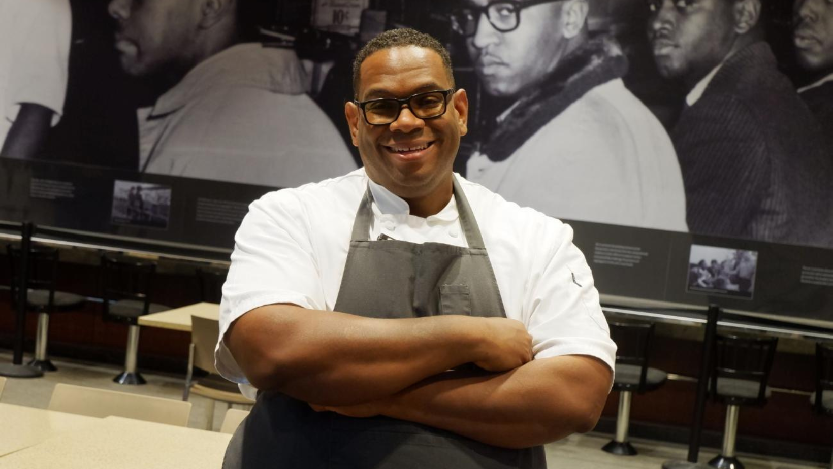 Chef Ramin Coles stands with his arms crossed, smiling widely. He wears a white chef jacket and a black apron.