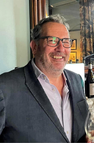 A photo of Andy woolgar smiling to the camera. He has rectangular glasses and wears a dark grey suit with a loosely buttoned button down underneath.