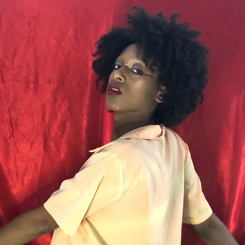 background is dark red satin. in the foreground a brown skinned nonbinary person wears a cream blouse, short cut afro, and red and black performance makeup