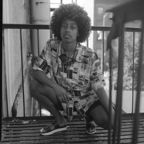 black and white image of a brown skinned non binary person kneeling on a fire escape. they are wearing shorts and a loose fitting blouse
